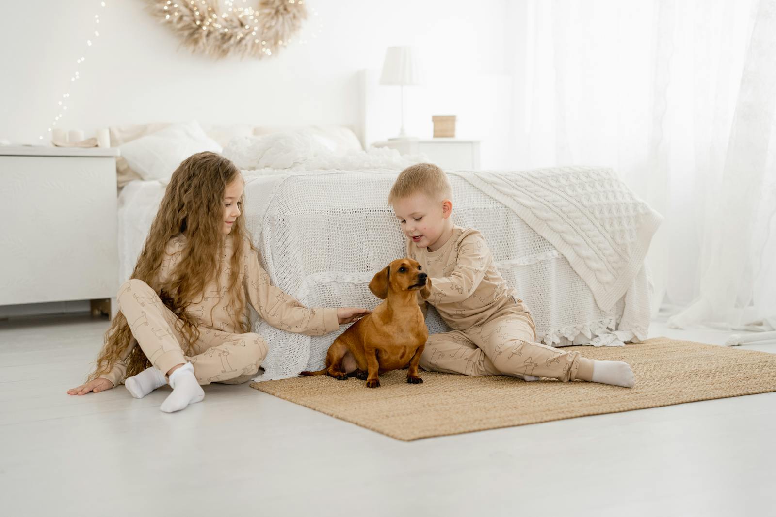 Two children in pajamas play with their dachshund dog in a bright, cozy bedroom.